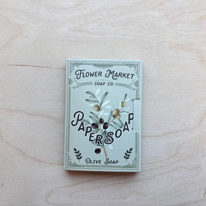 A square box of Paper Soap Olive Soap by Flower Market Soap Co. lies on a light wood grain surface.