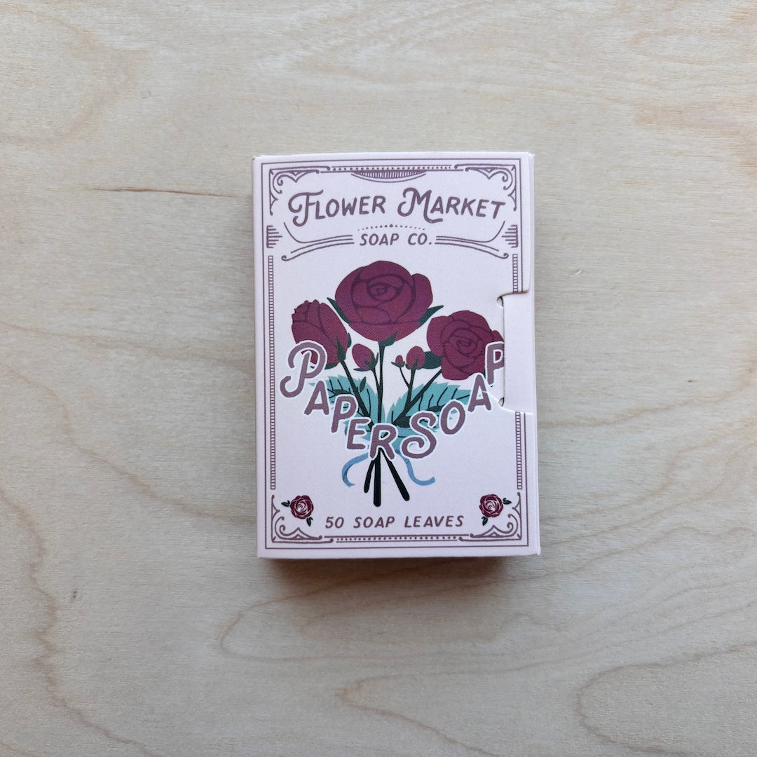 A boxed soap product labeled 'Flower Market Soap Co. Paper Soap' with an illustration of dark red roses and teal leaves, placed on a light wood surface.