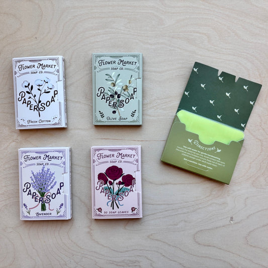 Four scented soap bars with illustrated paperboard boxes are arranged on a light wood surface, alongside a green envelope-style soap box. The soaps are labeled Flower Market soap bars, including Fresh Cotton, Olive Oil, Lavender, and Rosemary, with one box opened to reveal a bright green soap bar inside.