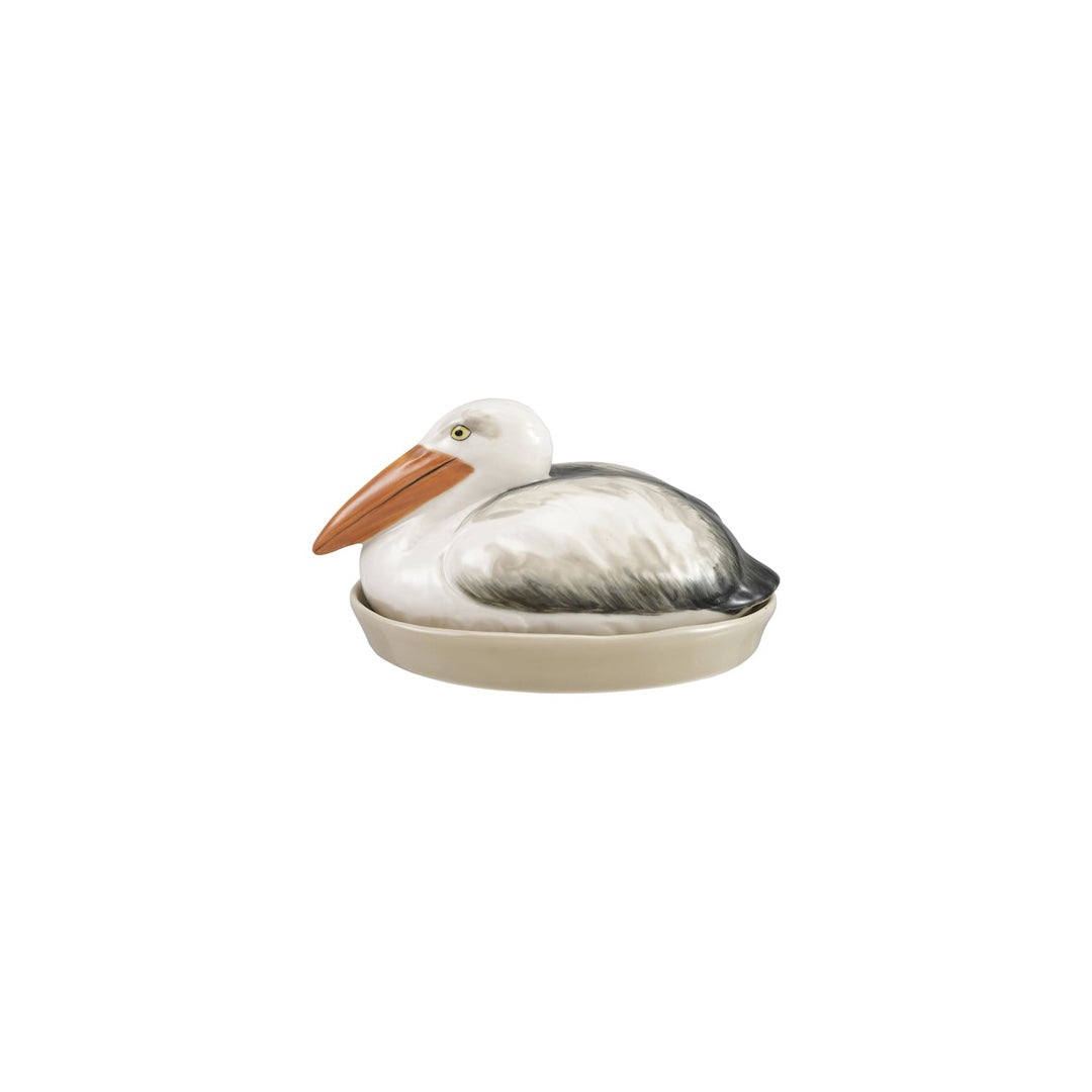 A butter dish that has a pelican figurine resting as a top in a shallow oval dish, featuring a white head and body, gray wings, and a peach-orange beak, set against a plain white background.