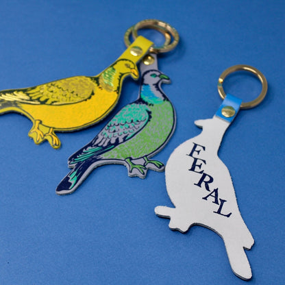 Three bird-shaped keychains on a blue background. The one on the right shows the back side of the keyring which has 'feral' written on it