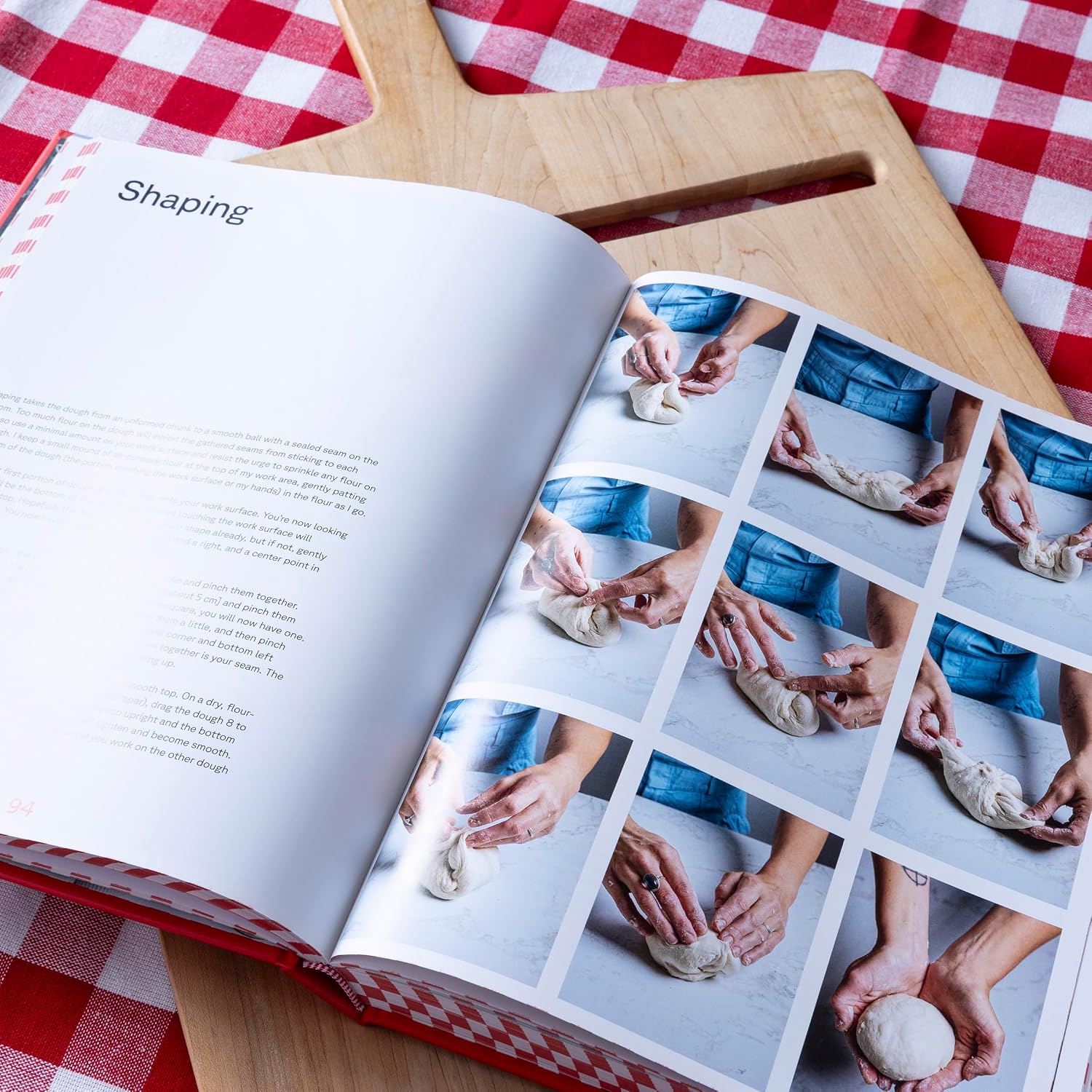 Open cookbook on a wooden cutting board with images of hands shaping dough, on a red and white checkered tablecloth.