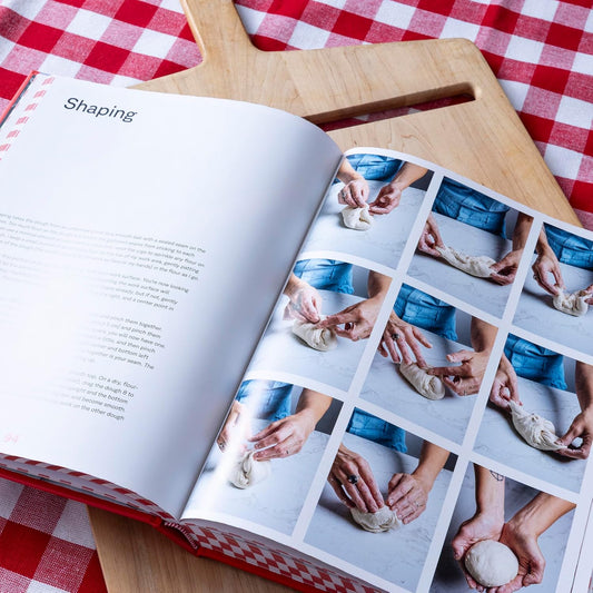 Open cookbook on a wooden cutting board with images of hands shaping dough, on a red and white checkered tablecloth.