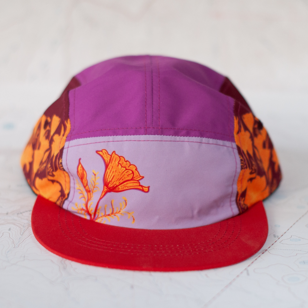 5 panel hat with a red brim and different shades of purple on each panel. Hat features poppy artwork