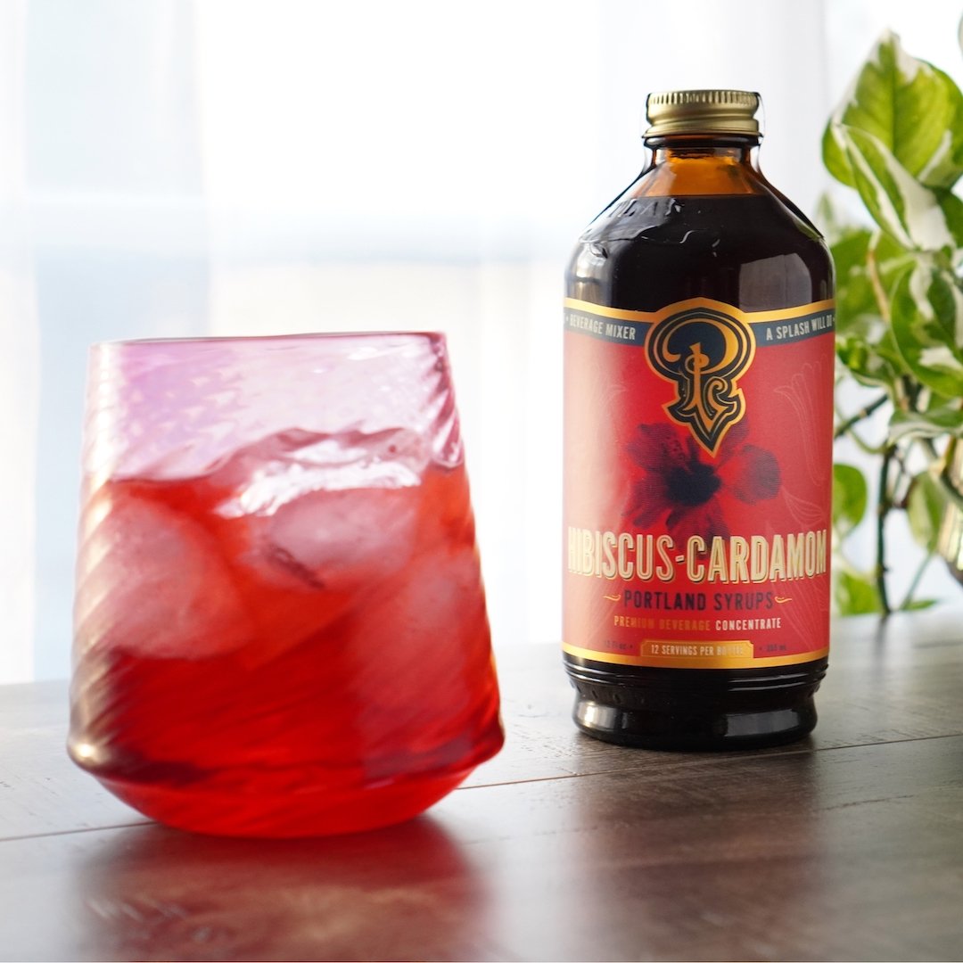 Bottle of hibiscus Cardamom syrup next to a glass with a red drink on a wooden surface.
