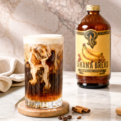 A bottle of Portland Syrups Banana Bread Syrup stands next to a glass of iced coffee with milk swirling in, set on a marble surface with cinnamon sticks and coffee beans.