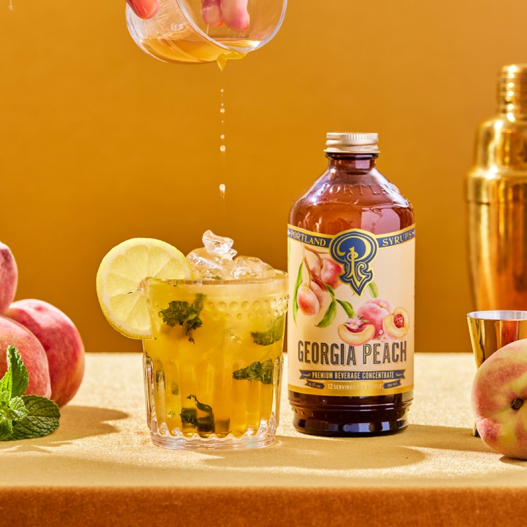 Georgia Peach syrup bottle with a cocktail being poured and peaches and mint leaves on a yellow background