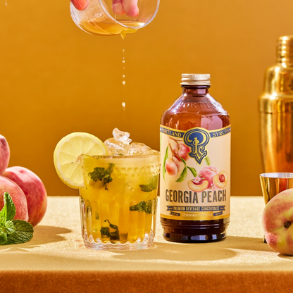 Georgia Peach syrup bottle with a cocktail being poured and peaches and mint leaves on a yellow background