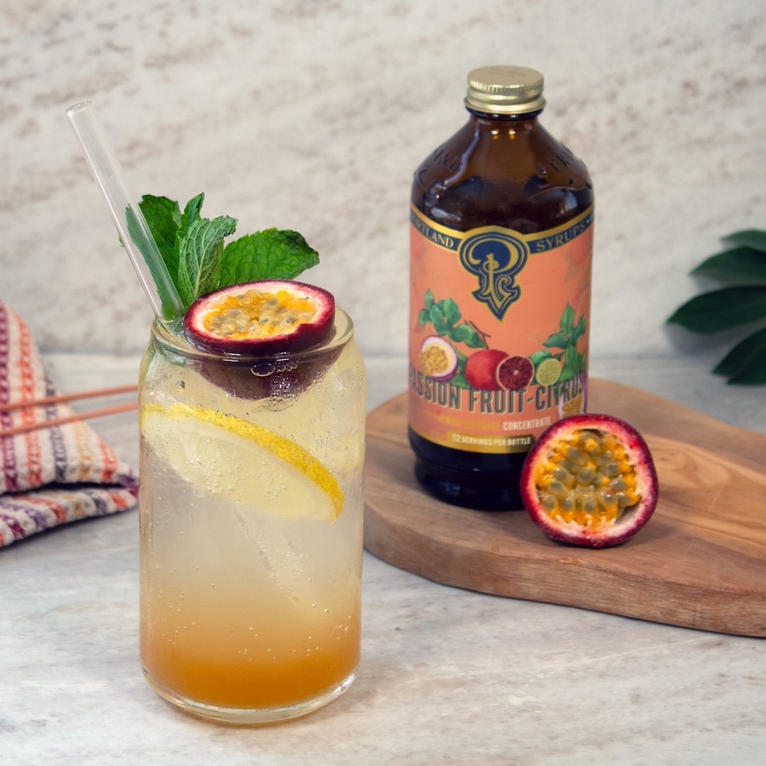 Cocktail with fruit garnish next to a bottle of passionfruit citrus syrup on a wooden board.
