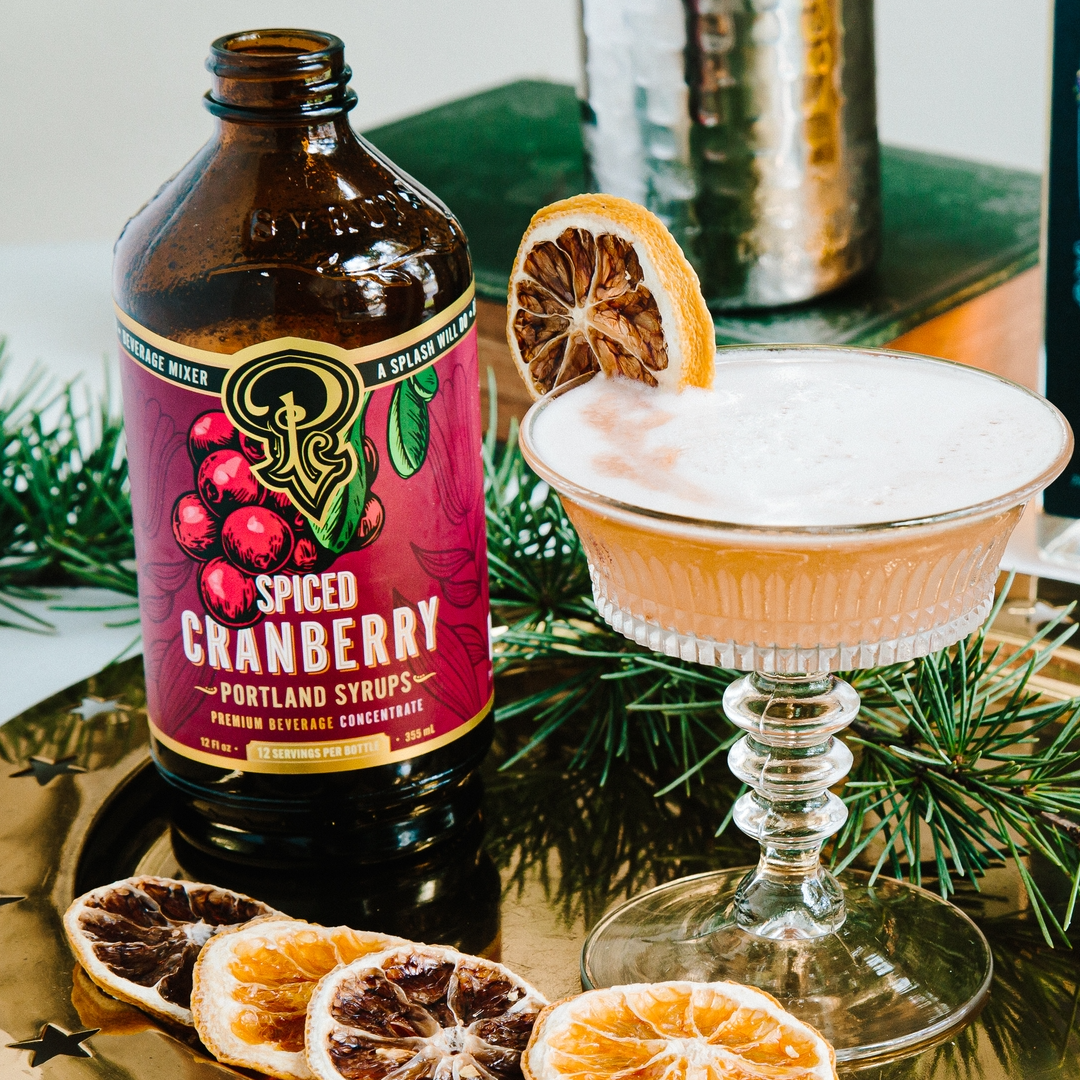 A bottle of Portland syrups Spiced Cranberry Portland Syrups next to a prepared cocktail in a coupe glass, garnished with a dried orange slice, set on a gold tray with dried orange slices and greenery.