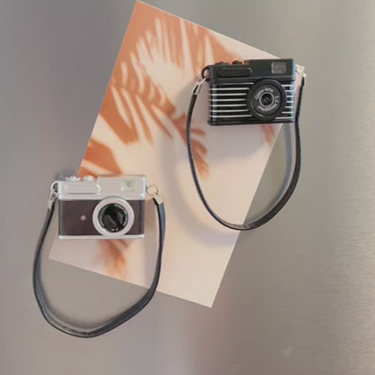 A video showing two miniature retro camera magnets on a fridge. A hand reaches out to show that pressing the button on top makes the flash on the camera work along with a flashing sound. The hand also turns the camera around to show the magnet on the back.