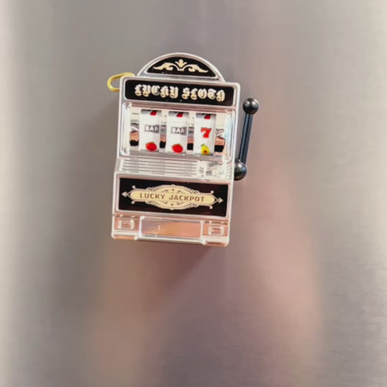 Video showing a mini slot machine magnet on a fridge. A hand reached out to pull the lever to show that the machine is interactive. The hand then pulls the magnet off the fridge to show the magnet on the back.