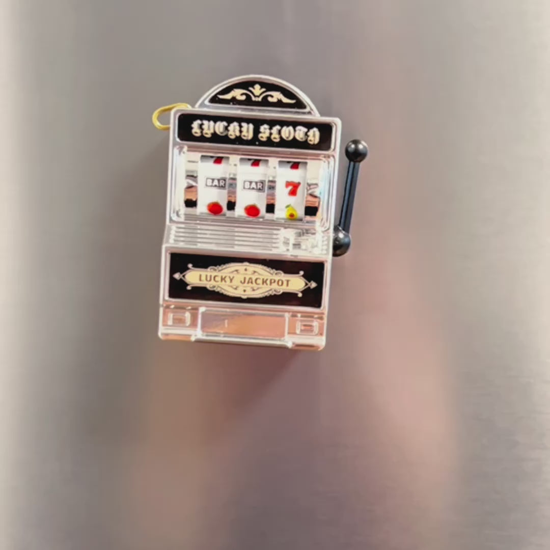 Video showing a mini slot machine magnet on a fridge. A hand reached out to pull the lever to show that the machine is interactive. The hand then pulls the magnet off the fridge to show the magnet on the back.
