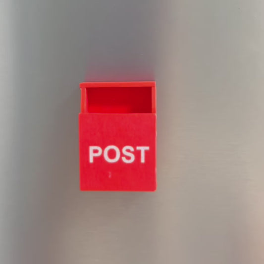 A video showing a small post box on a fridge. A post it note then comes into frame and a hand folds the post it and puts it into the box. The hand takes the post box off the fridge to show the magnet on the back 