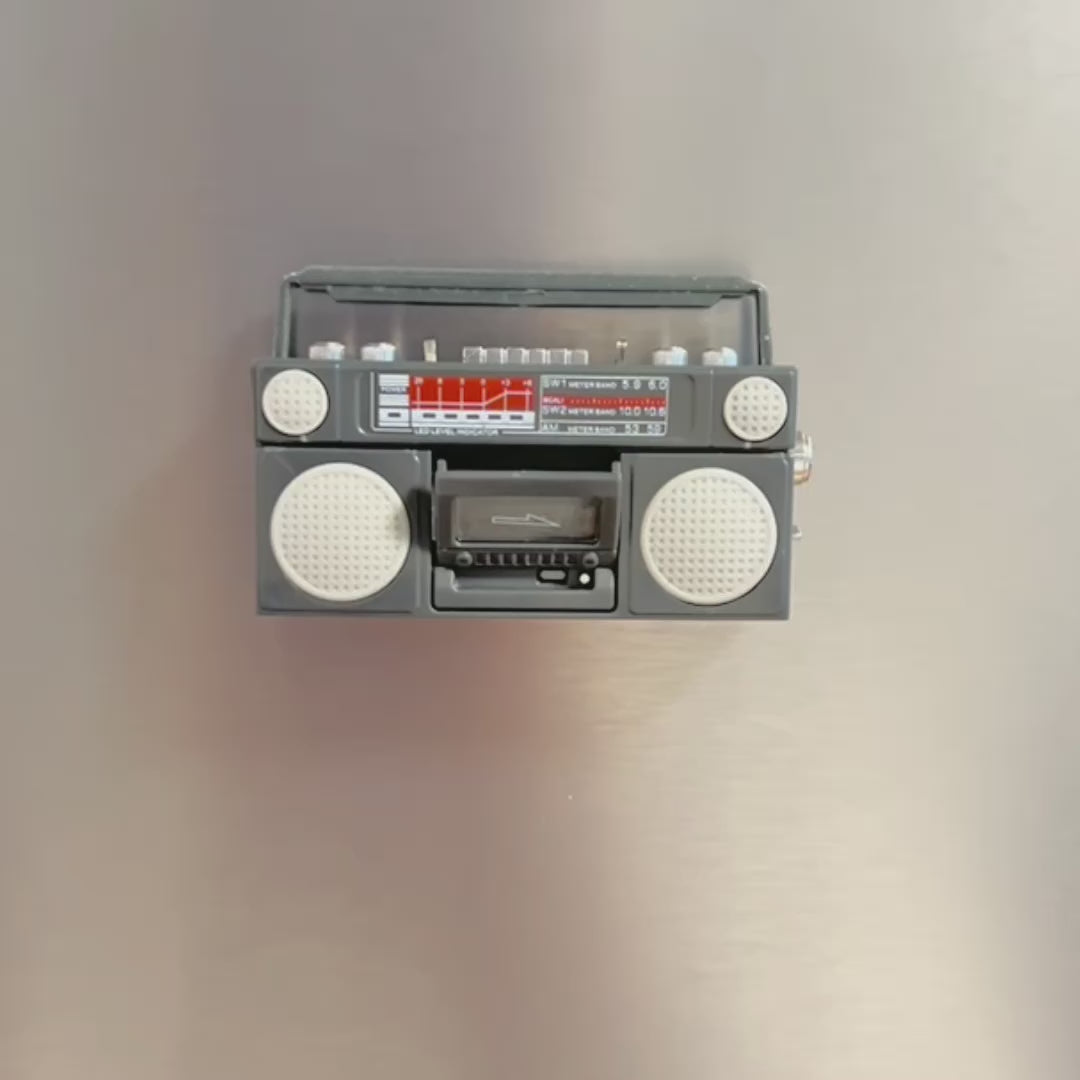 Video showing retro-style boombox-shaped refrigerator magnet on a fridge. A hand then reaches info frame to put a tape in the boombox then picks it up off the fridge to show the magnet on the back