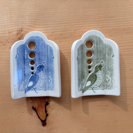 2 ceramic herb strippers. Both have 5 different sized holes to pull herbs through to remove the leaves. The one on the left has blue stripes and a quail at the bottom, the one on the right has green stripes and a quail on the bottom.