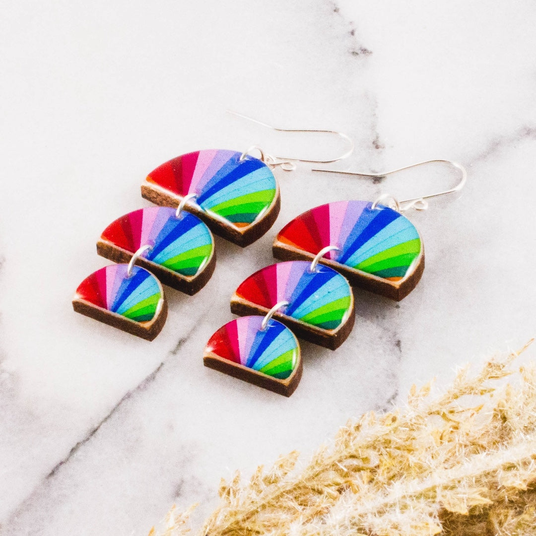 A pair of dangling earrings with colorful rainbow fan-shaped charms on a white marble surface with dried grass in the corner.
