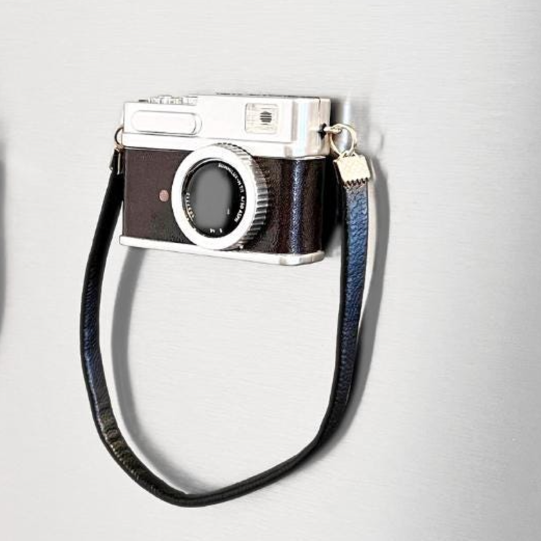 A novelty camera-shaped refrigerator magnet with a dark brown leather-look body, silver-tone metal details, and a black wrist strap, shown on a light gray textured background.