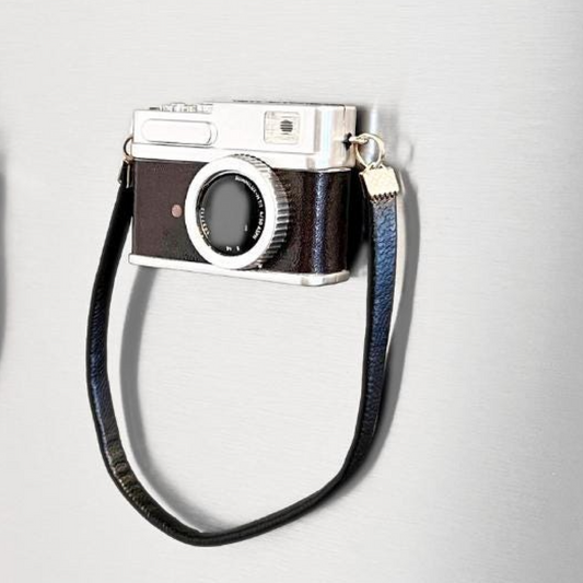 A novelty camera-shaped refrigerator magnet with a dark brown leather-look body, silver-tone metal details, and a black wrist strap, shown on a light gray textured background.