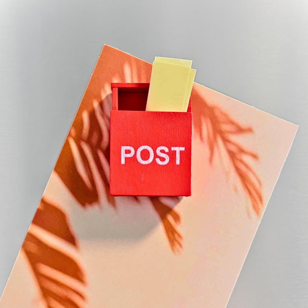 A small red post box with the word POST in white letters, holding two yellow sticky notes, placed on a peach-toned card with orange palm leaf shadows against a light gray background.