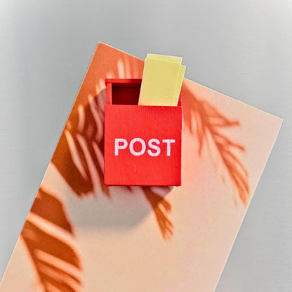 A small red post box with the word POST in white letters, holding two yellow sticky notes, placed on a peach-toned card with orange palm leaf shadows against a light gray background.