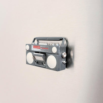 A retro-style boombox-shaped refrigerator magnet mounted on a brushed stainless steel refrigerator door.