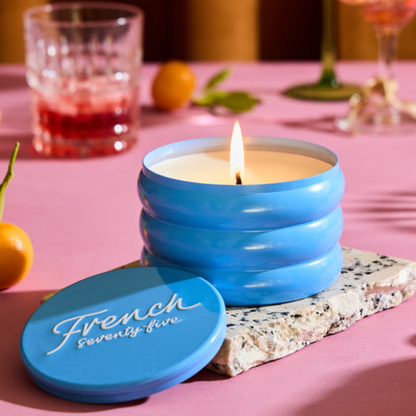 Blue candle in a yin container with 'French seventy-five' on the lid, surrounded by fruits and glasses on a pink surface.