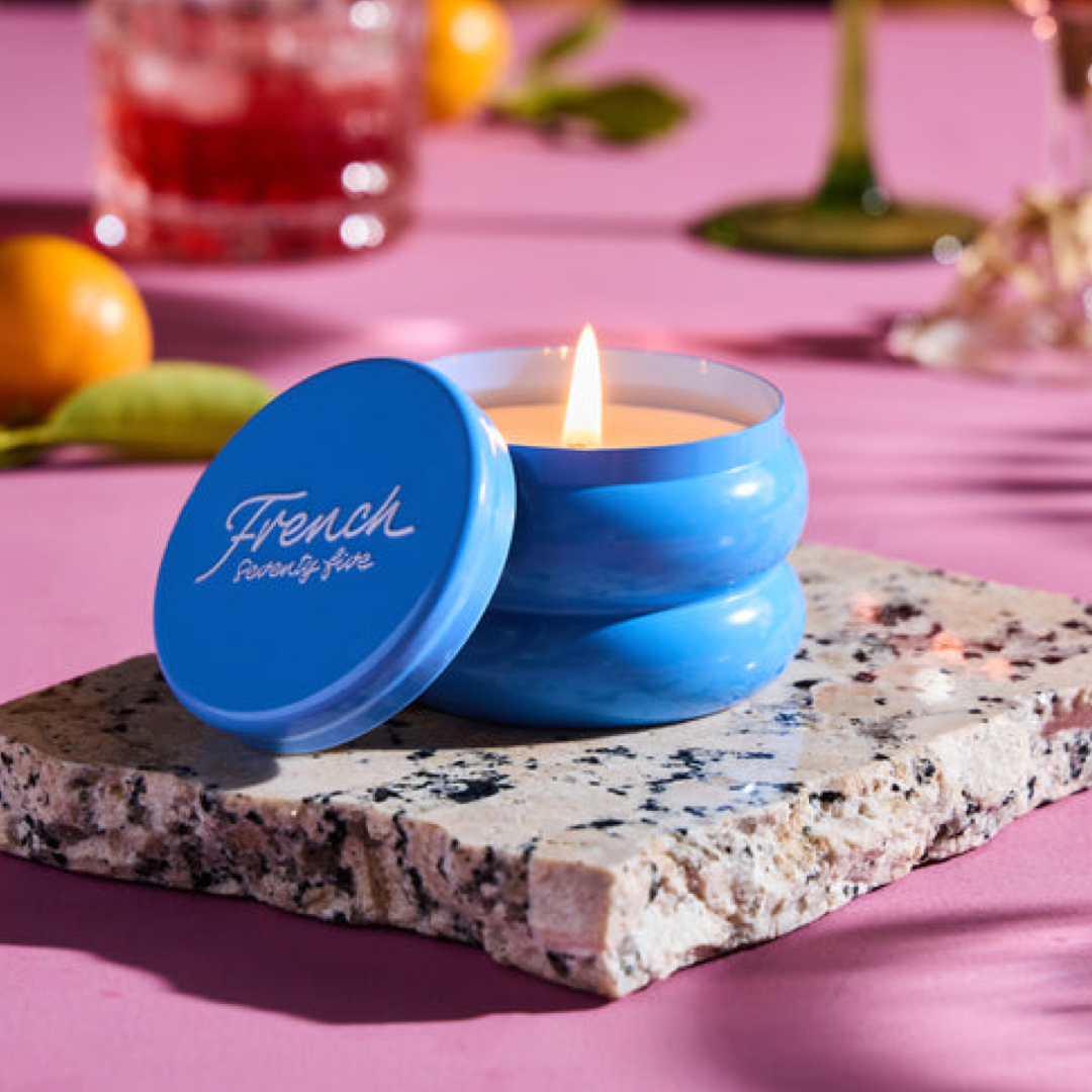 Blue candle with a lit wick on a pink surface, the lid has 'French seventy-five' on it.