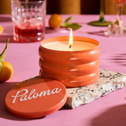 Candle with 'Paloma' branding on a pink surface with oranges and glasses in the background