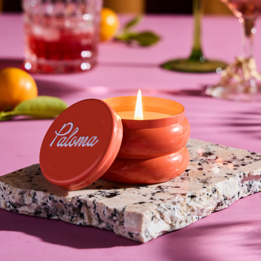 Orange candle in a tin with 'Paloma' branding on a pink tablecloth.