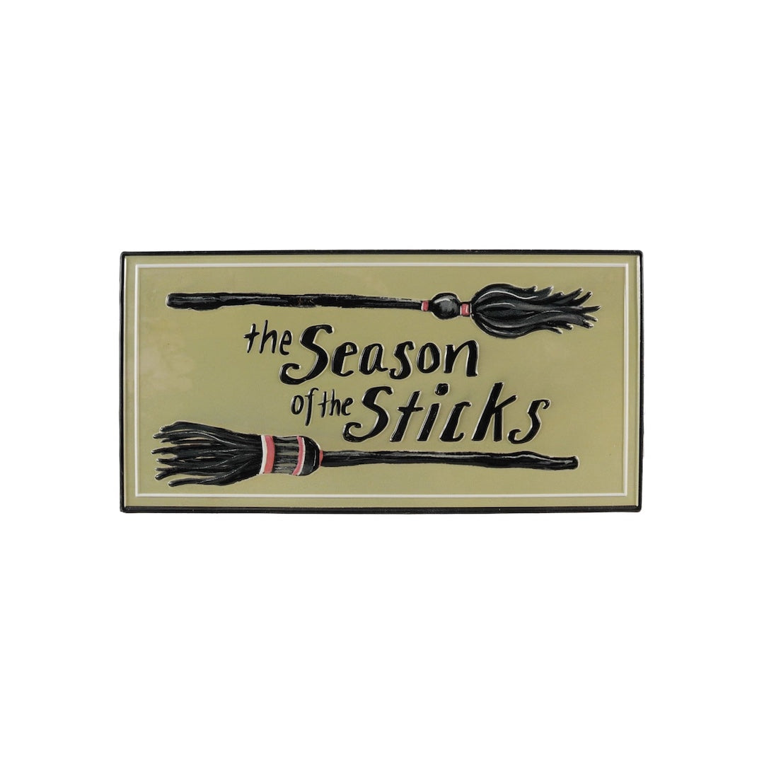 A rectangular decorative sign with a muted olive-beige background and black border, featuring two illustrated broomsticks and the phrase 'the Season of the Sticks' in black script.