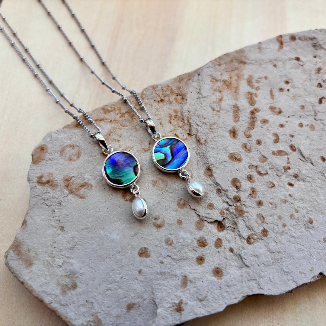 Two silver-toned beaded necklaces with oval abalone shell pendants and dangling pearl-like drops, displayed on a textured stone slab over a light wood surface.