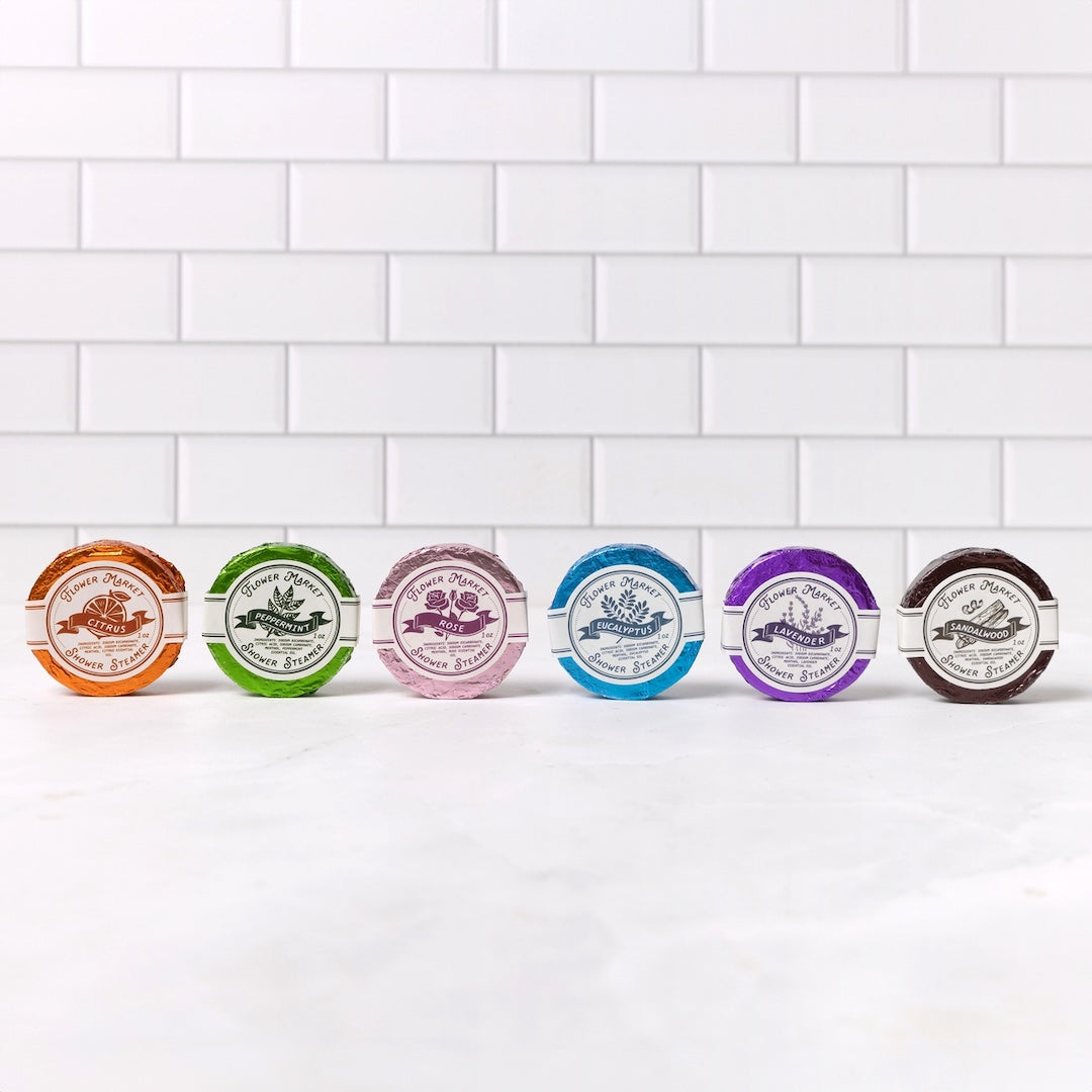 Six small round packaged items shower steamers lined up on a white marble surface against a white subway tile wall. Each package has a different colored rim and a white circular label with botanical-style illustrations and text.