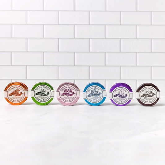 Six small round packaged items shower steamers lined up on a white marble surface against a white subway tile wall. Each package has a different colored rim and a white circular label with botanical-style illustrations and text.