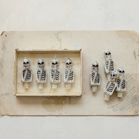 A set of novelty skeleton candles displayed on a textured beige background. The product includes 4 skeleton candles inside a small wooden tray on the left, and four skeleton candles standing outside the tray on the right.