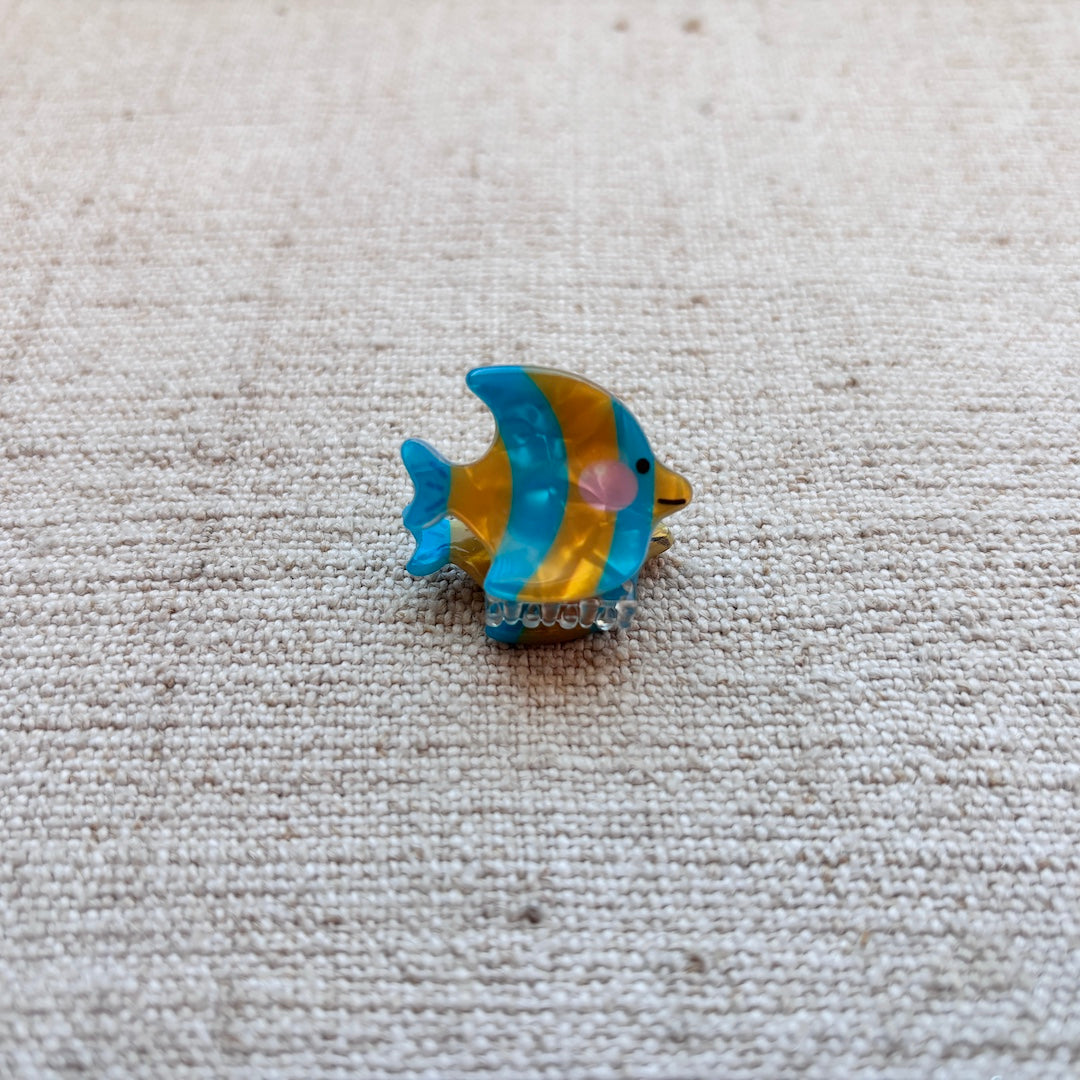 A small fish-shaped hair clip with blue and yellow stripes, a pink cheek, and a clear rhinestone base, placed on a textured beige fabric background.