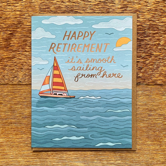 A square retirement greeting card featuring a hand-drawn sailboat on blue ocean waves with the text 'HAPPY RETIREMENT it's smooth sailing from here,' placed on a wooden surface.