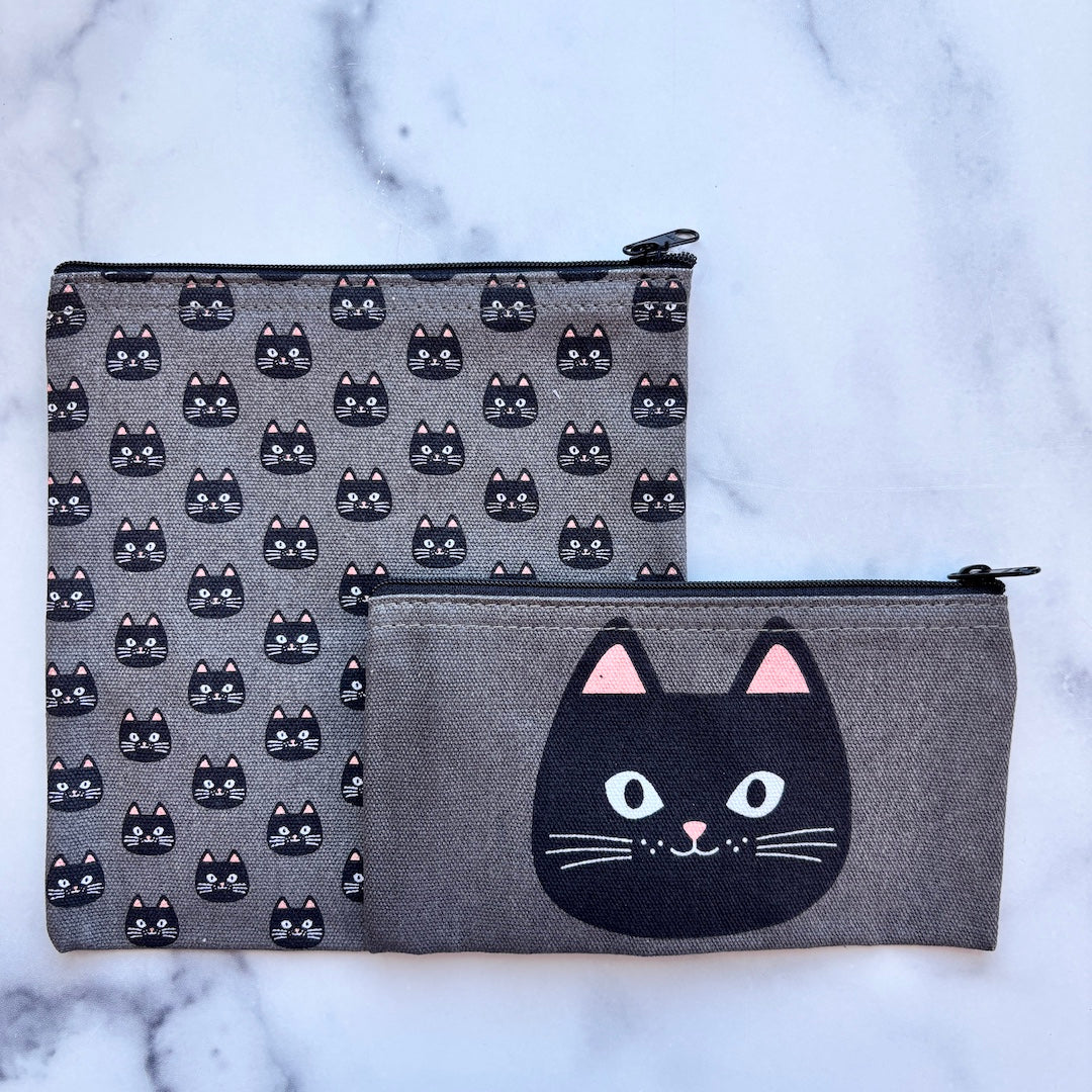 Two gray fabric zip pouches with black cat designs, displayed on a white marble surface. The larger pouch features a repeating pattern of black cat faces, and the smaller pouch features a large black cat face graphic on the front with pink inner ears.
