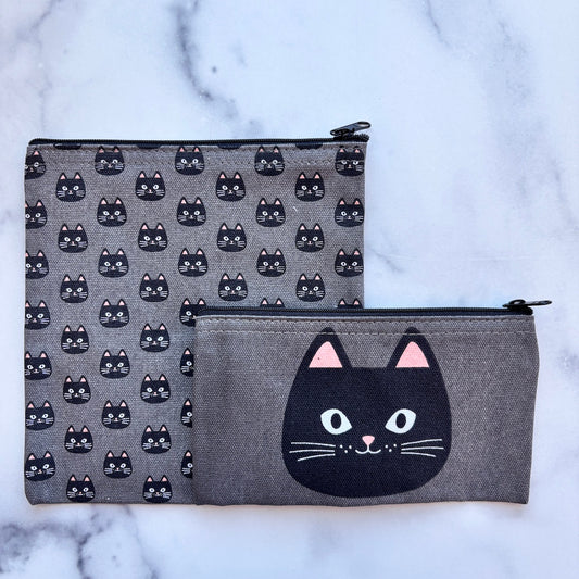 Two gray fabric zip pouches with black cat designs, displayed on a white marble surface. The larger pouch features a repeating pattern of black cat faces, and the smaller pouch features a large black cat face graphic on the front with pink inner ears.