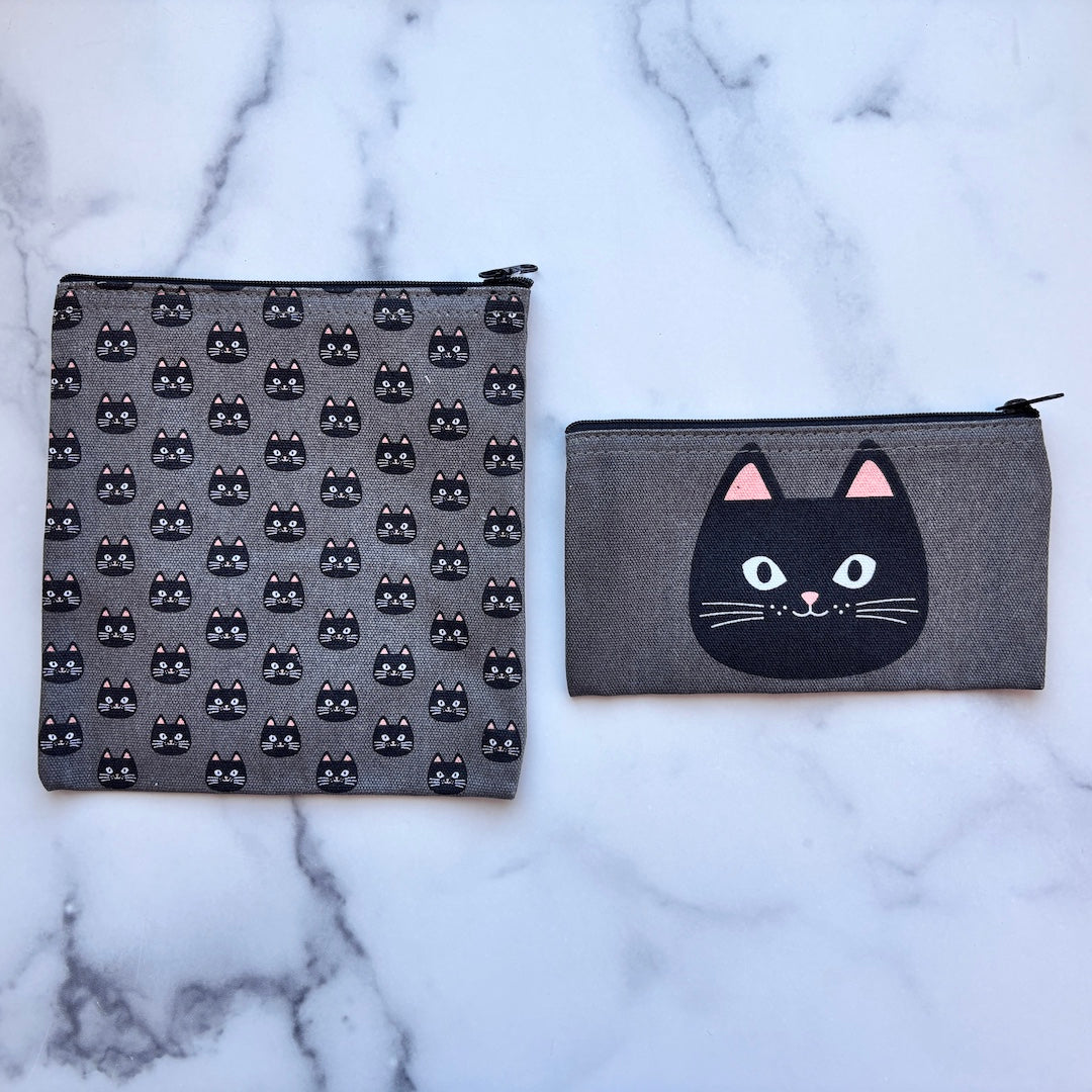 Two gray fabric zip pouches with black cat designs, displayed on a white marble surface. One pouch features a repeating pattern of small black cat faces, and the other features a large black cat face with pink ears on the front.