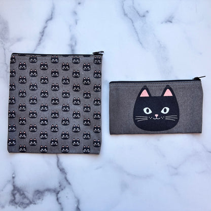 Two gray fabric zip pouches with black cat designs, displayed on a white marble surface. One pouch features a repeating pattern of small black cat faces, and the other features a large black cat face with pink ears on the front.