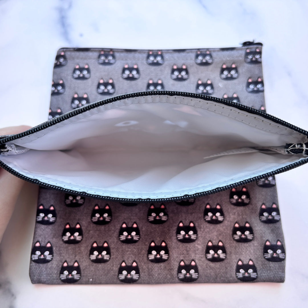 A zippered pouch with a gray fabric exterior printed with a repeating pattern of black cat faces with pink inner ears, shown open with a hand holding the left edge to display the white interior lining.