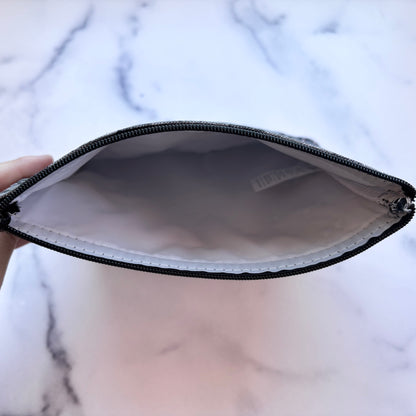 A close-up of a zippered pouch shown open, held by a hand against a white marble background. The pouch has a black zipper, a white interior lining, and a small white brand tag visible inside.