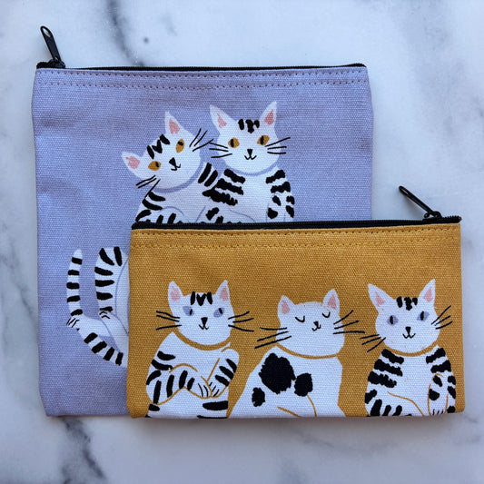 Two zippered fabric pouches with a black-and-white cat illustration, displayed on a light marble surface. The larger pouch has a lavender background, and the smaller pouch has a mustard yellow background.