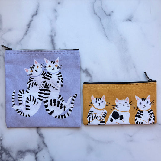 Two fabric zip pouches with black-and-white cat illustrations, displayed on a white marble surface. One pouch is light lavender, and the other is mustard yellow.