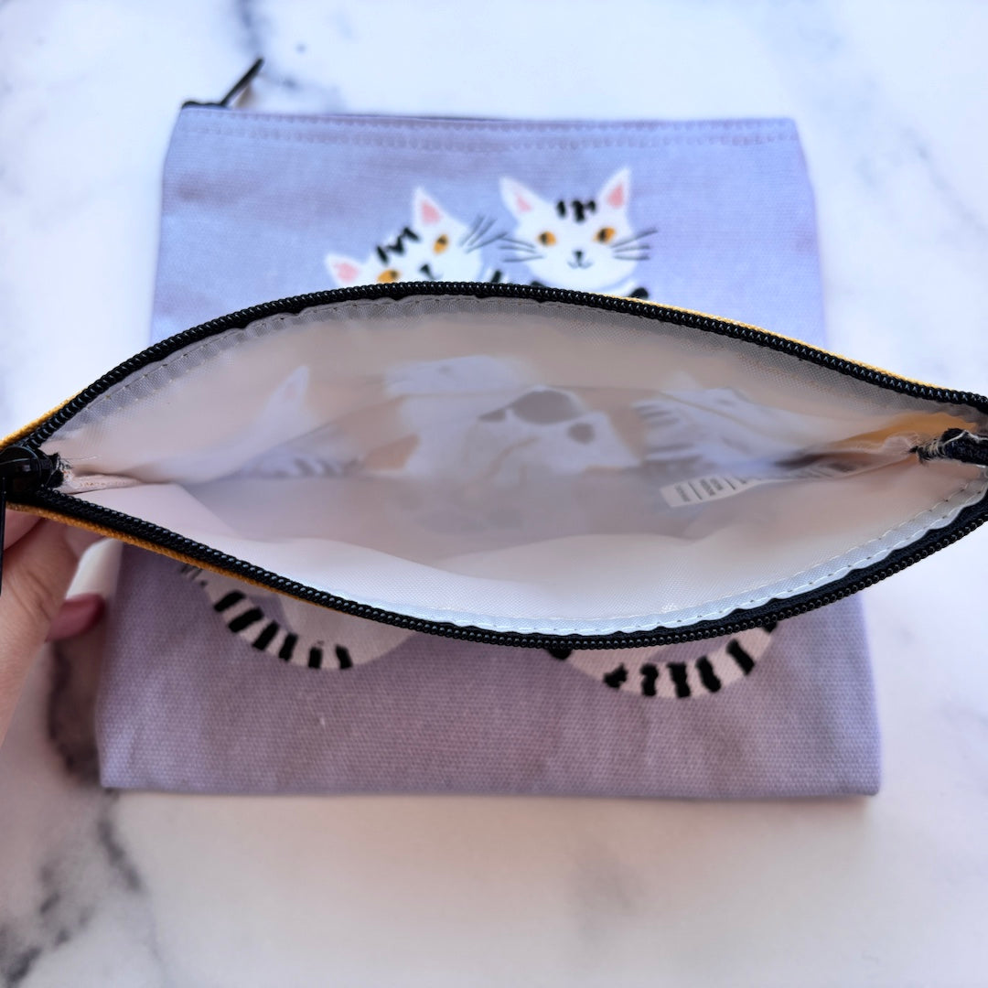 A lavender fabric zippered pouch with a cat face and striped cat-ear design, shown open with a hand holding one side to reveal the white lined interior. The pouch is placed on a white marble surface.