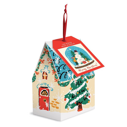 A Christmas-themed mini puzzle ornament shaped like a house, featuring festive illustrations of a snowman in a snow globe, a decorated Christmas tree, a red door, and a red hanging ribbon. A red tag on the roof reads '130 PIECE MINI PUZZLE' and 'Merry Christmas'.
