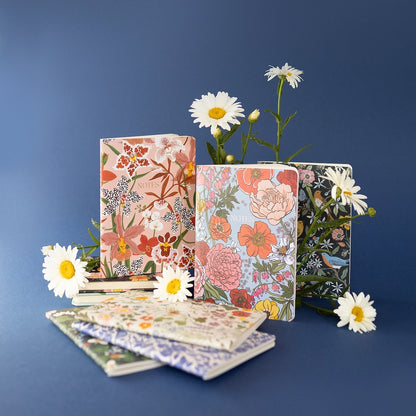 3 notebooks standing upright with different floral patterned covers surrounded by daisies. In the foreground is 3 notebooks laying flat.
