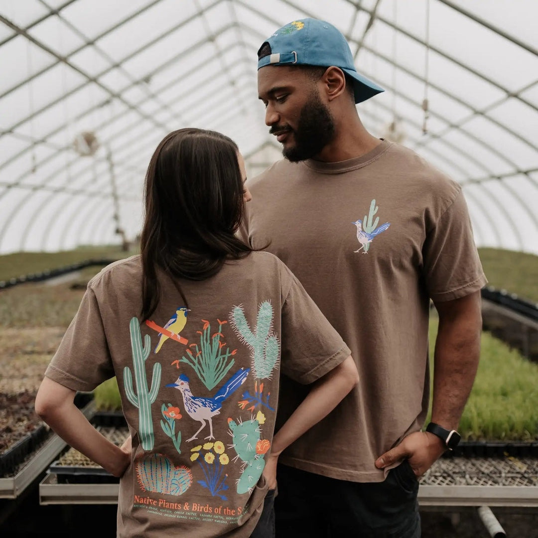 Two people wearing brown t-shirts with nature-themed designs in a greenhouse setting.
