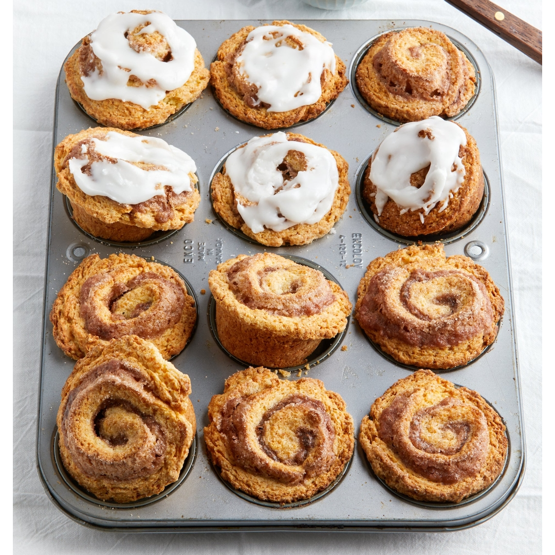 Twelve cinnamon rolls, including four topped with white icing, arranged in a metal 12-cup muffin tin on a light fabric surface.